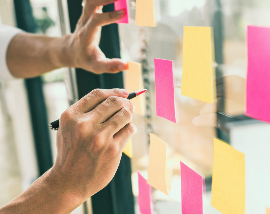 Verschiedenfarbige Post-it Notes hängen an einer Glasfront und werden mit einem Stift beschrieben